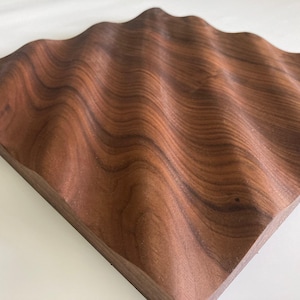 May include: A square wooden tray with a wavy surface. The tray is made of dark brown wood with visible grain patterns. The edges are clean and the design is minimalist. The tray is approximately 25 cm x 25 cm.