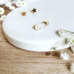 May include: A set of gold-toned earrings featuring a crescent moon and star design. The earrings are displayed on a white surface with white flowers.