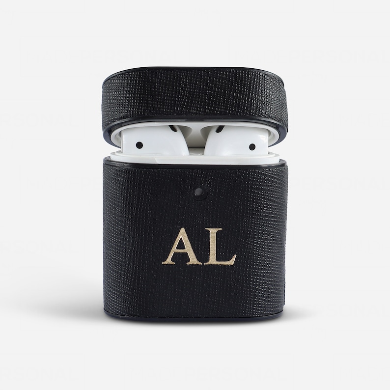 May include: A black textured protective case for wireless earbuds with a hinged lid. The front of the case features the gold embossed letters AL. Inside the open case, two white wireless earbuds are visible, showing their smooth, rounded design.