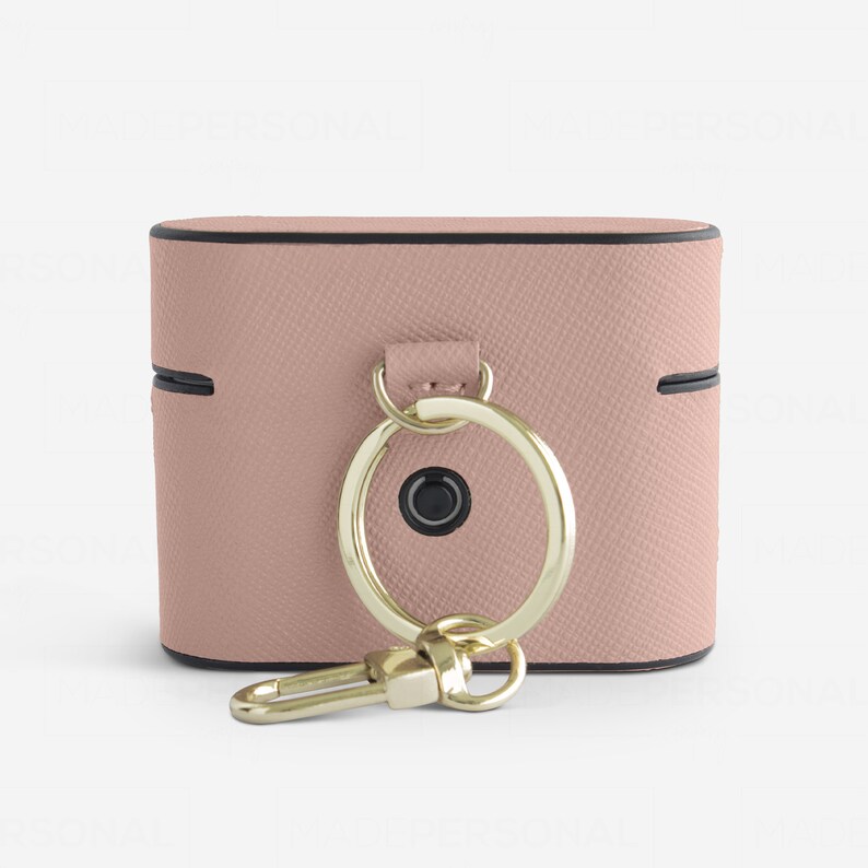 May include: A dusty pink textured protective case for wireless earbuds featuring a gold-toned metal ring and a matching gold-toned lobster clasp attachment. The case has a black trim along the edges and a small circular black button on the front.