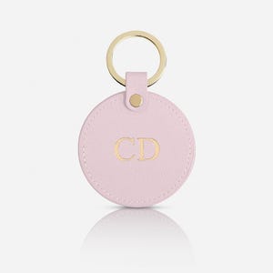 May include: A circular light pink keychain featuring a textured finish and the gold embossed letters CD in the center. The round tag is attached to a gold-toned metal split ring by a small matching pink leather tab secured with a gold metal rivet.
