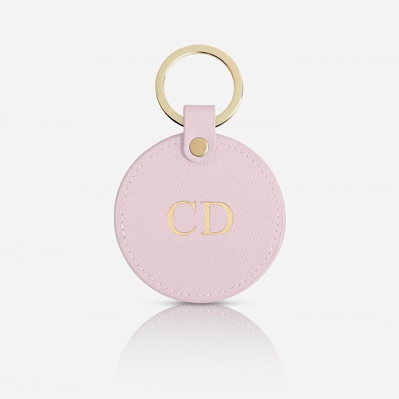 May include: A circular light pink keychain featuring a textured finish and the gold embossed letters CD in the center. The round tag is attached to a gold-toned metal split ring by a small matching pink leather tab secured with a gold metal rivet.
