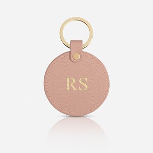 May include: A circular, dusty pink keychain featuring a textured surface and gold-toned hardware. The center of the round tag is embossed with the gold-colored capital letters RS. A gold metal ring is attached to the top via a matching pink tab.