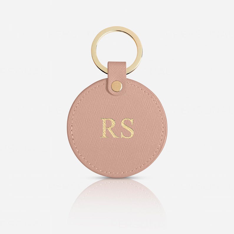 May include: A circular, dusty pink keychain featuring a textured surface and gold-toned hardware. The center of the round tag is embossed with the gold-colored capital letters RS. A gold metal ring is attached to the top via a matching pink tab.