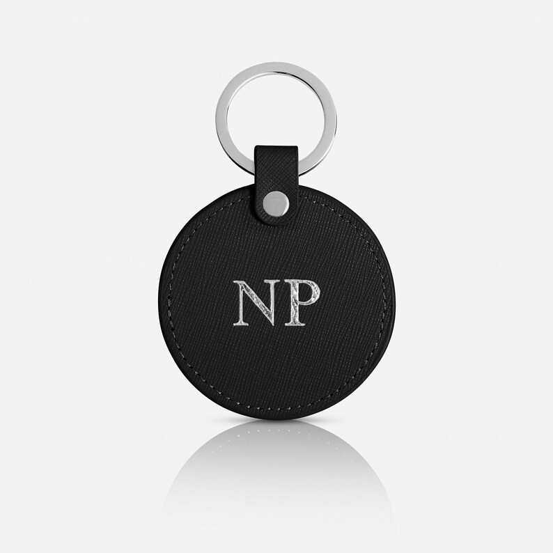 May include: A circular black keychain featuring a textured surface and the silver embossed letters NP in the center. The keychain is attached to a silver metal split ring by a small black tab secured with a single silver-colored metal rivet.