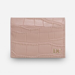May include: A dusty rose card holder featuring a textured crocodile skin pattern. The rectangular accessory has visible stitching along the edges and the gold-colored capital letters DC are embossed in the bottom right corner of the front panel.