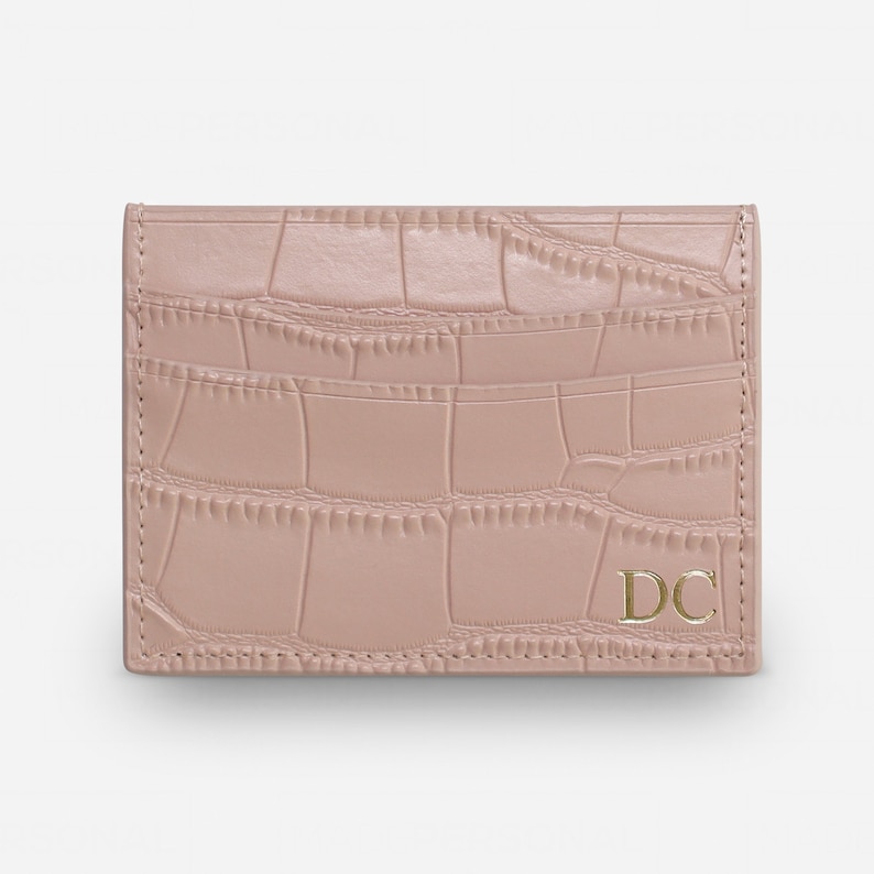 May include: A dusty rose card holder featuring a textured crocodile skin pattern. The rectangular accessory has visible stitching along the edges and the gold-colored capital letters DC are embossed in the bottom right corner of the front panel.