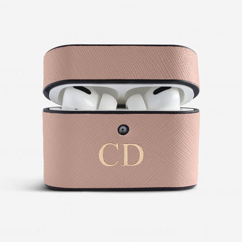 May include: A dusty pink textured protective case for wireless earbuds, featuring a black trim and the gold embossed letters CD on the front. The case is open, revealing white wireless earbuds inside, and includes a small circular cutout for a light.