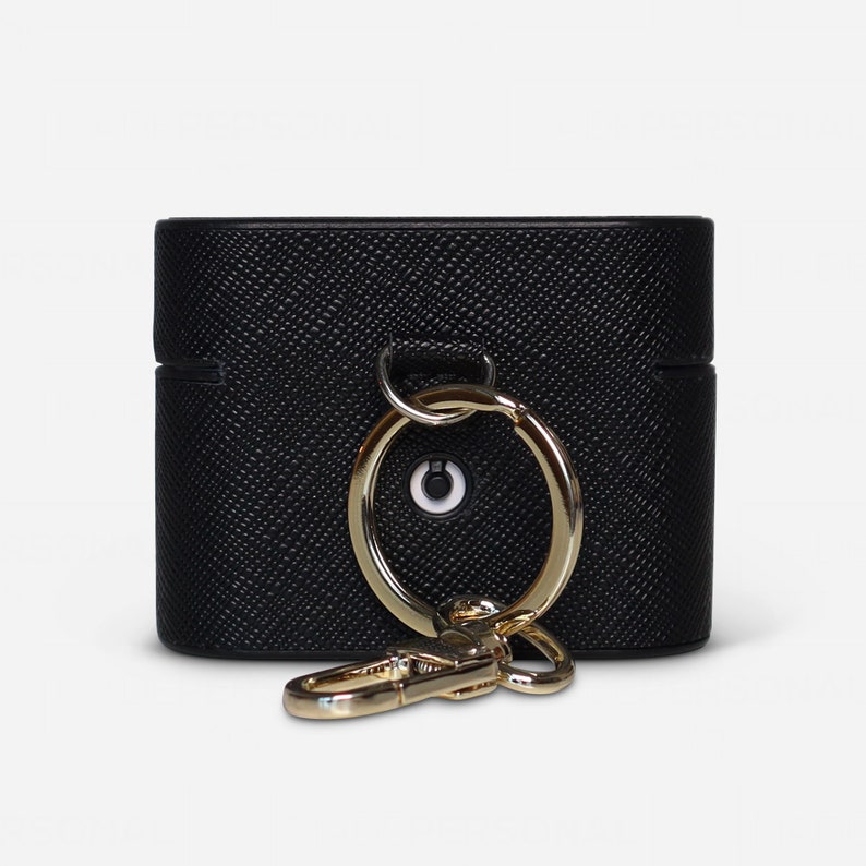 May include: A black textured protective case for wireless earbuds featuring a gold-toned metal ring and a matching lobster clasp attachment. The case has a horizontal seam across the middle and a small circular cutout revealing a white indicator light.