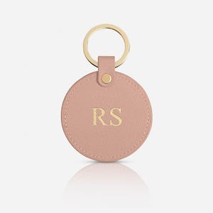 May include: A circular, dusty pink keychain featuring a textured surface and gold-toned hardware. The center of the round tag is embossed with the gold-colored capital letters RS. A gold metal ring is attached to the top via a matching pink tab.