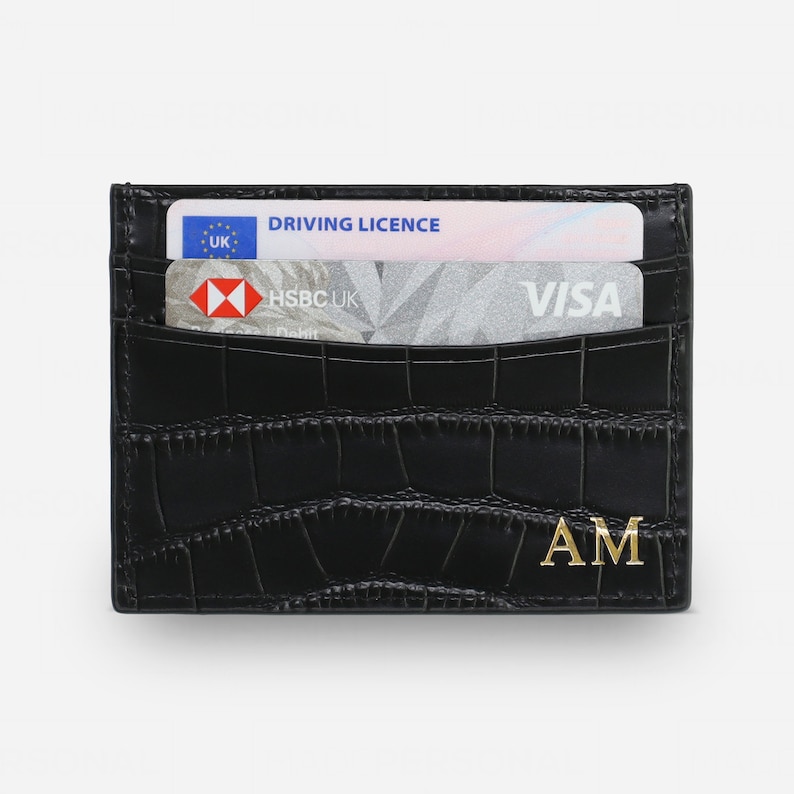 May include: A black card holder with a crocodile-embossed texture and gold-colored initials AM in the bottom right corner. The card holder contains a UK driving license and an HSBC UK Visa debit card inserted into the top slot.