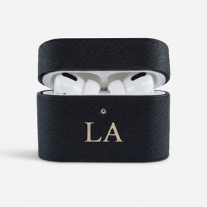 May include: A black textured protective case for wireless earbuds, shown in an open position with white earbuds inside. The front of the case features the gold-coloured letters LA centred below a small circular indicator light on the lid.