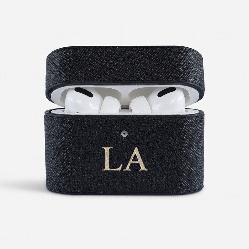 May include: A black textured protective case for wireless earbuds, shown in an open position with white earbuds inside. The front of the case features the gold-coloured letters LA centred below a small circular indicator light on the lid.
