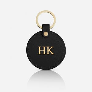 May include: A circular black leather keychain featuring a textured surface and the gold embossed letters HK in the center. The keychain is attached to a gold-toned metal split ring by a small leather tab secured with a single gold-colored metal rivet.