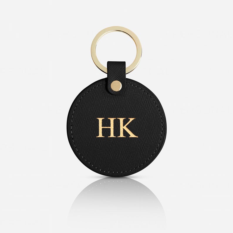 May include: A circular black leather keychain featuring a textured surface and the gold embossed letters HK in the center. The keychain is attached to a gold-toned metal split ring by a small leather tab secured with a single gold-colored metal rivet.
