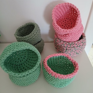 Op de afbeelding: Een verzameling handgemaakte gehaakte manden in verschillende maten en kleuren. De manden zijn in tinten groen, roze en grijs. Sommige hebben contrasterende randen. Deze decoratieve manden zijn ideaal voor het organiseren van kleine items of als woondecoratie.