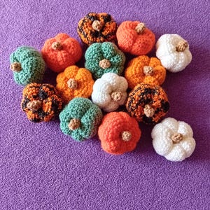 May include: A selection of crocheted pumpkins in shades of orange, green, white, and black with orange detailing. Each pumpkin features a small, brown stem. The pumpkins are displayed on a purple surface.