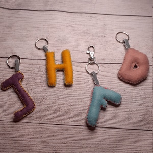 Könnte beinhalten: Vier Schlüsselanhänger aus Filz in verschiedenen Farben: ein lila "J", ein gelbes "H", ein blaues "F" und ein rosa "D".