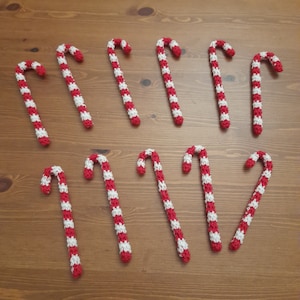 Puede incluir: Nueve bastones de caramelo de ganchillo a rayas rojas y blancas, colocados sobre una superficie de madera. Estos bastones curvados son una decoración festiva clásica, ideal para Navidad.