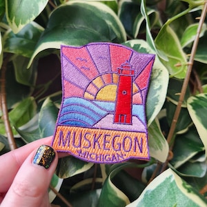 May include: Embroidered patch featuring a red lighthouse, a setting sun, and the text "Muskegon Michigan." The design includes blue waves, birds, and a purple sky. The patch is rectangular with a curved top edge.