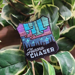 May include: Embroidered patch featuring a shield design with a colorful depiction of the aurora borealis. The patch includes shades of purple, teal, and blue, with the words "Aurora Chaser" in white script.