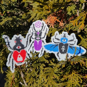 May include: Three colorful beetle stickers with wings and an eye design. The beetles are purple, blue, and red with black accents. The stickers are on a green leafy background.