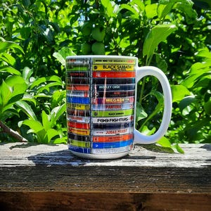 May include: White ceramic mug featuring a design of stacked cassette tapes with band names like Black Sabbath, Led Zeppelin, and Foo Fighters. The mug has a white handle and is set against a backdrop of green foliage.