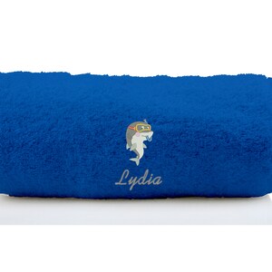 K&ouml;nnte beinhalten: Ein blaues, zusammengerolltes Handtuch mit einem grauen, gestickten Delfin, der eine Taucherbrille tr&auml;gt, und dem Namen "Lydia", der darunter gestickt ist.