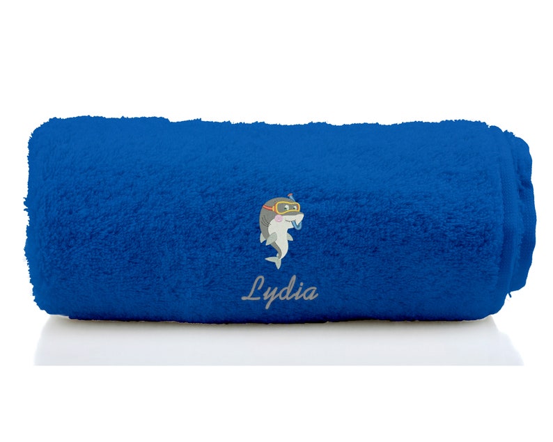 K&ouml;nnte beinhalten: Ein blaues, zusammengerolltes Handtuch mit einem grauen, gestickten Delfin, der eine Taucherbrille tr&auml;gt, und dem Namen "Lydia", der darunter gestickt ist.