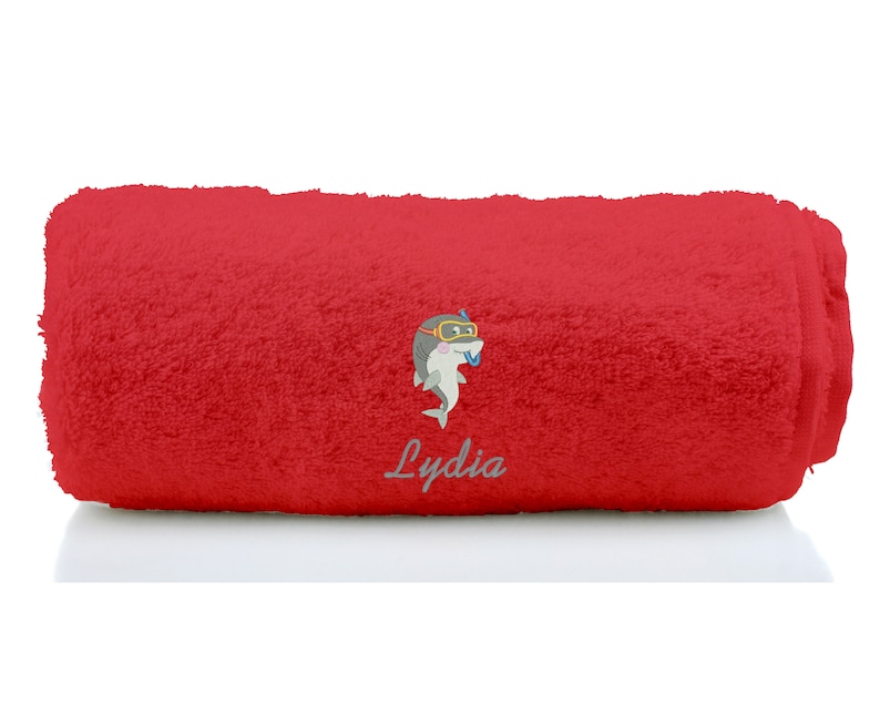 K&ouml;nnte beinhalten: Rotes Badetuch mit einem Cartoon-Hai, der einen Schnorchel und eine Taucherbrille tr&auml;gt. Das Handtuch ist aufgerollt und der Name "Lydia" ist auf das Handtuch gestickt.
