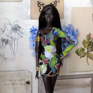 May include: A dark-skinned fashion doll in a one-shoulder mini dress with a floral print in green, white, and blue. The doll has long black hair and gold arm cuffs. The background features floral artwork and a butterfly image.