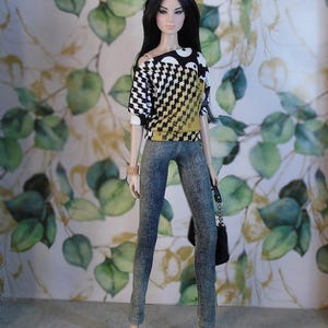 May include: A doll dressed in a patterned top with black, white, and yellow elements, paired with denim-style trousers and beige heels. The doll has long black hair and a gold headband. A black handbag with a silver chain is also visible.