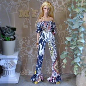 May include: A fashion doll with long blonde hair wearing an off-the-shoulder jumpsuit featuring a white, navy blue, yellow, and pink geometric pattern. The jumpsuit has long sleeves, a cinched waist, and ruffled hems. A gold necklace is worn.