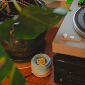 May include: A blue ceramic wax warmer with a honeycomb-shaped wax melt on top. The warmer is sitting on a wooden surface next to a black stereo system and a houseplant.