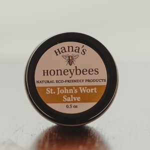 Könnte beinhalten: Eine kleine, runde Blechdose mit schwarzem Deckel und einem Etikett mit der Aufschrift "Hana's Honeybees Natural Eco-Friendly Products St. John's Wort Salve 0.5 oz". Das Etikett hat einen braunen Hintergrund mit einem gelben Streifen und einem kleinen Bild einer Biene.