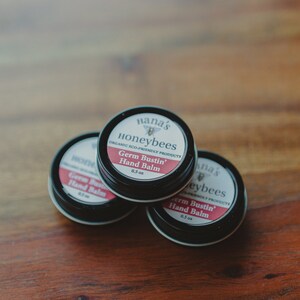 May include: Three small black tins of hand balm stacked on a wooden surface. The label on the top tin reads "Nana's Honeybees Organic Eco-Friendly Products Germ Bustin' Hand Balm 0.3 oz."