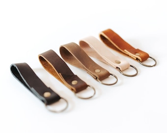 May include: A set of six leather keychains in various shades of brown, tan, and black. Each keychain features a simple, rectangular leather strap with a metal ring.