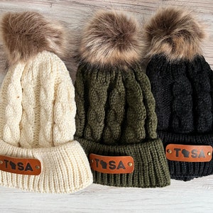 Puede incluir: Tres gorros de punto trenzado en crema, verde oliva y negro, cada uno rematado con un pompón de piel sintética marrón. Un parche de cuero marrón con las letras "TSA" y un contorno estatal está cosido en la parte delantera de cada gorro.