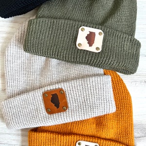 Op de afbeelding: Drie gebreide mutsen in verschillende kleuren: grijs, groen en mosterdgeel. Elke muts heeft een leren patch met de omtrek van de staat Illinois.