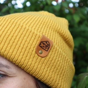 Peut inclure: Bonnet en tricot jaune moutarde avec un design côtelé. Un petit patch en cuir marron avec un motif de fromage est fixé à l'avant. Le bonnet est fait d'une matière douce et chaude, idéale pour l'hiver.