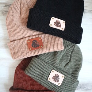 May include: Four knitted beanies in different colors, each with a leather patch featuring a leaf design. The beanies are stacked on top of each other, with the black beanie on top.