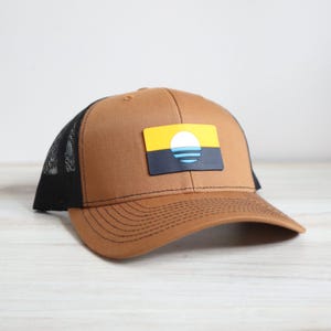 May include: A brown and black trucker hat with a black mesh back. The hat features a yellow, blue, and black patch with a stylized sun and waves design.