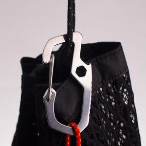 Op de afbeelding: Een zilveren karabijnhaak met een flesopener bevestigd aan een zwarte netzak met een rood en geel koord.