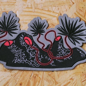 Puede incluir: Parche bordado con un diseño en negro y gris. El parche presenta un paisaje negro con detalles blancos, acentos rojos y tres formas grises similares a palmeras. El parche tiene un borde gris y es probablemente para ropa o accesorios.