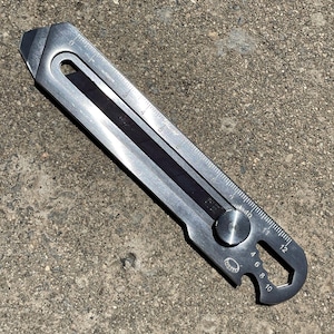 Puede incluir: Una multiherramienta plateada con regla, cuchilla y llave. La herramienta tiene una cuchilla negra y un botón de ajuste circular. La regla está marcada en centímetros.