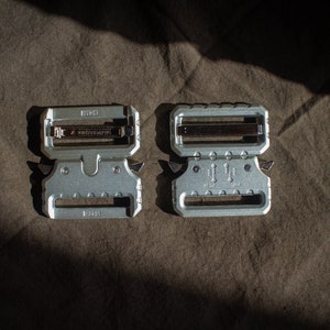 May include: Two silver metal belt buckles with a textured surface. The buckle on the left has the text "HK" and "2055" on it. The buckle on the right has the text "HK" and "2055" on it.