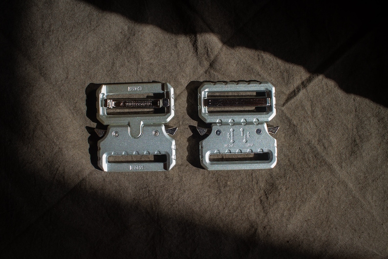 May include: Two silver metal belt buckles with a textured surface. The buckle on the left has the text "HK" and "2055" on it. The buckle on the right has the text "HK" and "2055" on it.