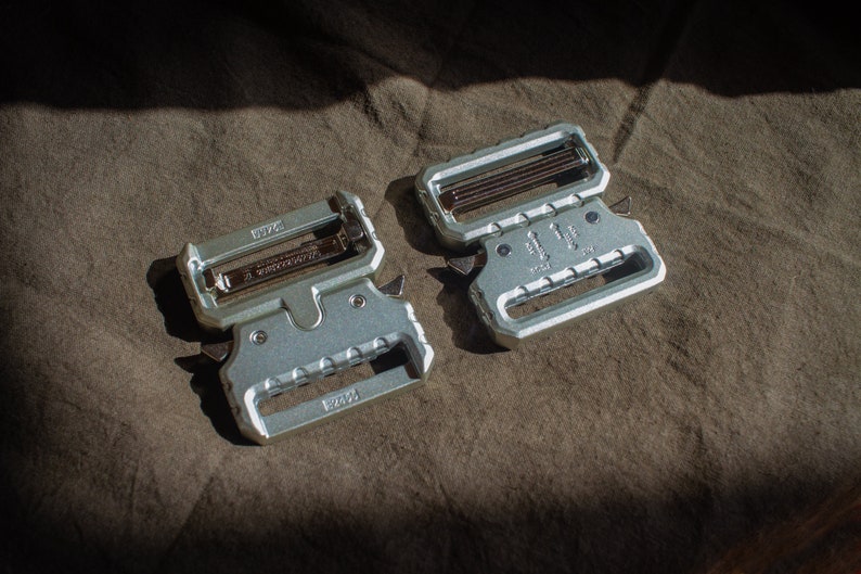 May include: Two silver metal belt buckles with a textured surface. The buckles have a silver metal pin and a black metal spring. The buckles are labelled "1.75" and "1.25".
