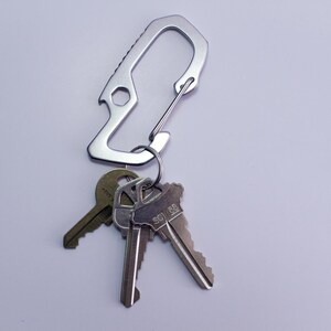 Op de afbeelding: Een zilveren sleutelhanger met een flesopener en een karabijnhaak. De sleutelhanger heeft drie sleutels eraan bevestigd.