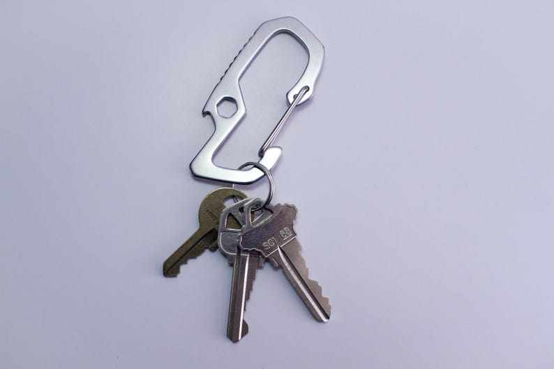 Op de afbeelding: Een zilveren sleutelhanger met een flesopener en een karabijnhaak. De sleutelhanger heeft drie sleutels eraan bevestigd.